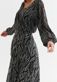 Robe noire à motifs de feuilles blanches, manches longues transparentes, taille froncée et bas fluide. Le tissu texturé ajoute de la profondeur au design.