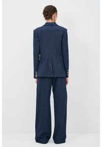 Costume bleu marine ajusté avec un blazer cintré orné de boutons dorés et un pantalon à jambes larges. Le tissu présente une texture subtile avec un design structuré.