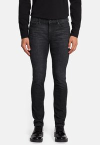 Jean en denim noir slim fit avec une finition légèrement décolorée, design à cinq poches et rivets en métal. Porté avec des chaussures noires à lacets.