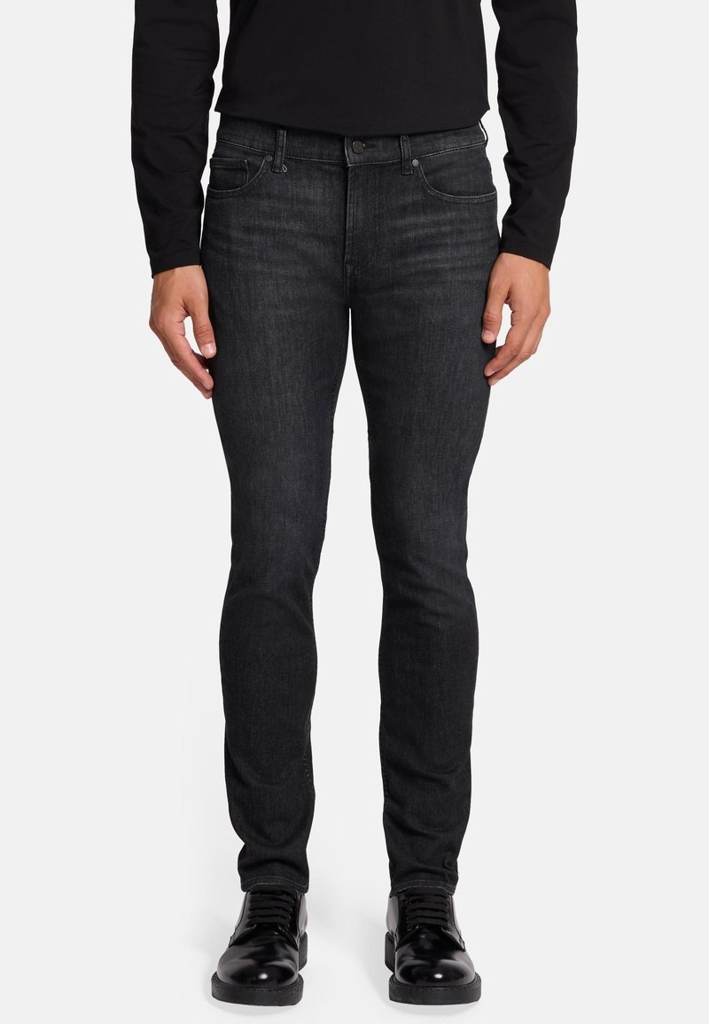 Jean en denim noir slim fit avec une finition légèrement décolorée, design à cinq poches et rivets en métal. Porté avec des chaussures noires à lacets.