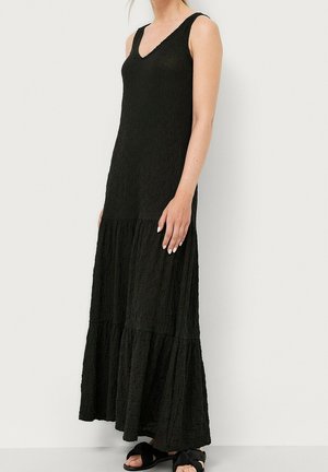 Femme portant une robe longue noire texturée sans manches avec un col en V, debout devant un fond clair uni, portant des sandales noires.
