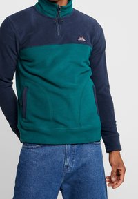 Pullover in pile con mezza zip, maniche blu scuro e corpo verde acqua. Include una patch con logo e due tasche laterali. Tessuto testurizzato.