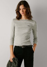 Camisola cinza de manga longa com um decote redondo e um logótipo subtil, combinada com calças pretas e uma camisa xadrez pendurada no braço.