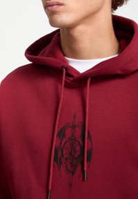 Sudadera color burdeos hecha de un tejido suave, que presenta un gráfico negro de una calavera, una espada y alas de murciélago en el frente. Cuerdas de ajuste con puntas negras.