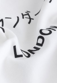 Tissu blanc avec texte partiel imprimé en noir comprenant "LONDON" et des caractères japonais, montré en gros plan avec des plis visibles.