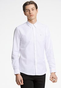 Camisa blanca de botón hecha de algodón con mangas largas, un cuello clásico y un sutil logo bordado en el pecho. Corte entallado, textura suave.