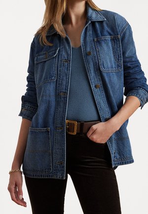 Persoon die een blauw geribd topje draagt, een blauwe spijkerjas met grote zakken, donkere broek en een bruine riem met een gegraveerde gesp.