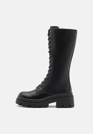 Sorte lærknæhøje støvler med en kraftig sål. Har snøringer foran og en glat overflade, hvilket giver et slankt og robust design.