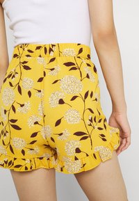 Geel bloemen shorts van een structuurstof, met witte en bruine bloempatronen, een gerimpelde zoom en een elastische tailleband.