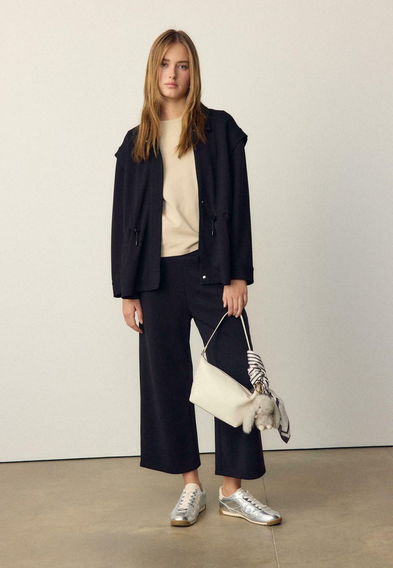 Jeune femme en veste et pantalon noirs, haut beige, baskets argentées, tenant un sac à main blanc avec foulard et porte-clés en peluche.