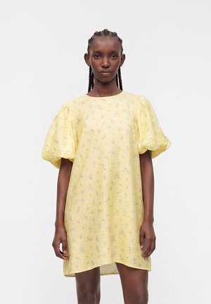 Frau mit geflochtenem Haar, die ein locker sitzendes gelbes Kleid mit Puffärmeln und kleinem Blumenmuster trägt, vor einem schlichten Hintergrund stehend.