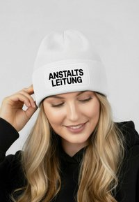 White knitted beanie with a rectangular black patch displaying the text "ANSTALTSLEITUNG." Soft texture, rounded top seam, and snug fit.