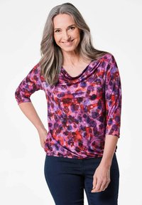 Lila und rote gemusterte Bluse mit einem Schalkragen und Dreiviertelärmeln, aus einem weichen, fließenden Stoff. Getragen mit dunkelblauen Jeans.