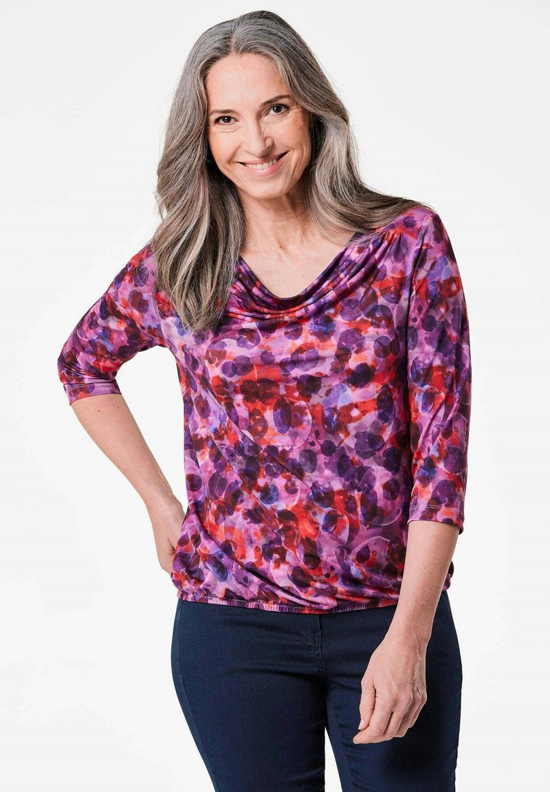 Lila und rote gemusterte Bluse mit einem Schalkragen und Dreiviertelärmeln, aus einem weichen, fließenden Stoff. Getragen mit dunkelblauen Jeans.