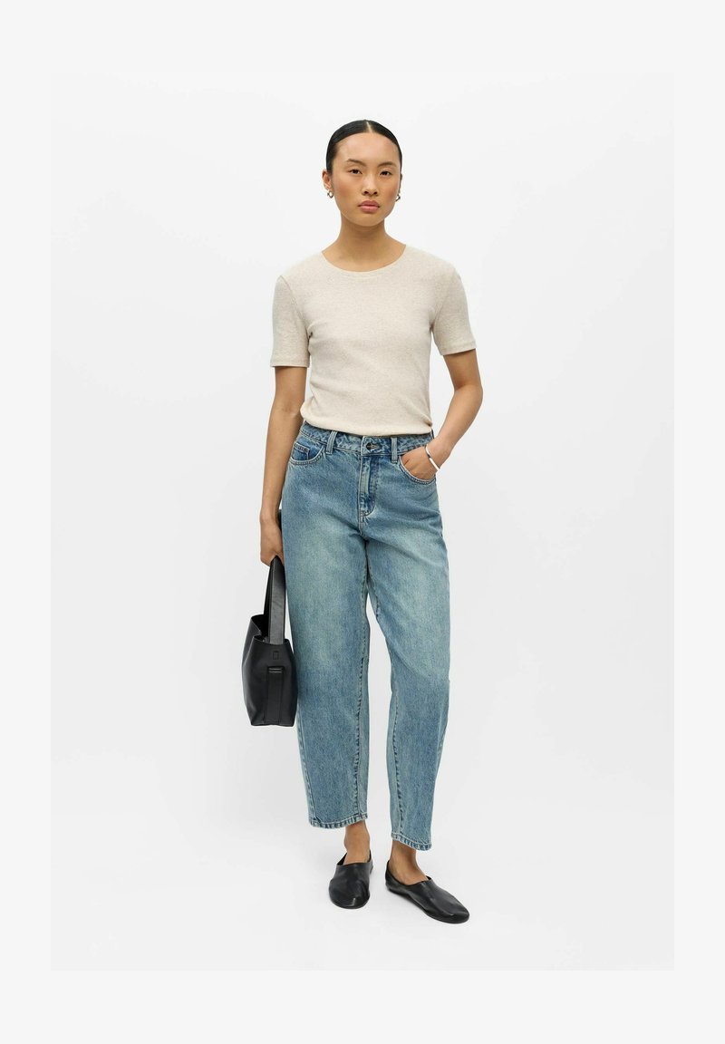 Femme debout, main dans la poche, portant un haut beige à manches courtes, un jean bleu, des chaussures noires sans lacets, et tenant un sac à main noir.