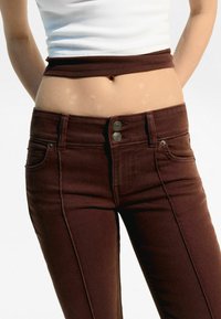 Jeans de mezclilla marrón con un ajuste slim, cintura alta y cierre de dos botones. Presenta bolsillos frontales y detalles de costura sutiles.