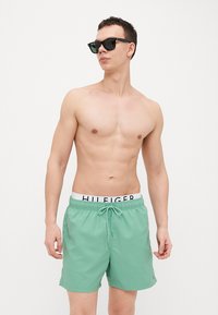 Shorts de bain verts avec une taille élastique, dotés d'un cordon de serrage et d'une bande avec logo. Le tissu semble lisse et léger.