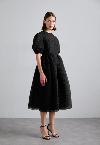 Black puff-sleeve dress with a textured pattern, cinched waist, and flowing skirt. Worn with black strappy heels.
