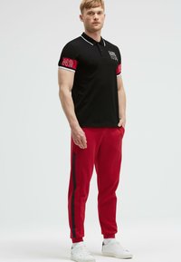 Polo nero con maniche a contrasto rosse, caratterizzato da bordi bianchi e logo stampato. Abbinato a pantaloni jogging rossi e scarpe da ginnastica bianche, creando un look sportivo.
