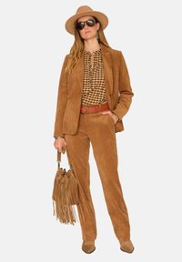 Un costume en velours côtelé marron avec une finition texturée, associé à un chemisier à carreaux. Les accessoires incluent un chapeau à large bord et un sac à franges.