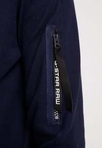 Marinblå, texturerad tröja med en zippered ficka på vänster ärm, med en svart dragflik och tryckt logotyp "G-STAR RAW."