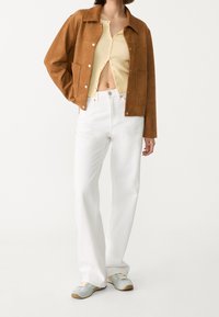 Brun ruskindsjakke over en gul ribstrikket top, kombineret med hvide høj-taljede bukser og lyseblå sneakers. Har trykknapper og lommer.