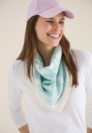 Femme souriante portant une casquette violet clair, un haut blanc et une écharpe à motifs bleu clair autour du cou, sur un fond neutre.