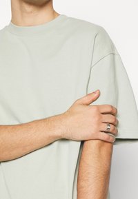 T-shirt vert clair en coton à manches courtes, avec un col rond. Une bague en argent orne la main visible reposant sur le bras.