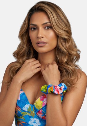 Kobieta o falowanych, jasnobrązowych włosach, ubrana w niebieską bluzkę w kwiaty i z kolorową, kwiatową gumką na nadgarstku, na jasnym tle.