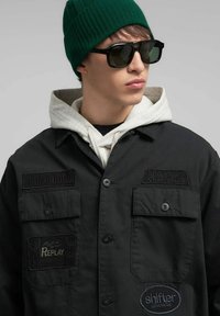 Chaqueta negra con botones al frente y bolsillos delanteros, decorada con varios parches bordados. Llevada sobre una sudadera con capucha gris, acompañada de gafas de sol y un gorro verde.