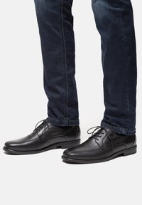 Scarpe eleganti in pelle nera con punta arrotondata e lacci, abbinate a jeans in denim blu scuro, mostrando una texture liscia e un design minimale.