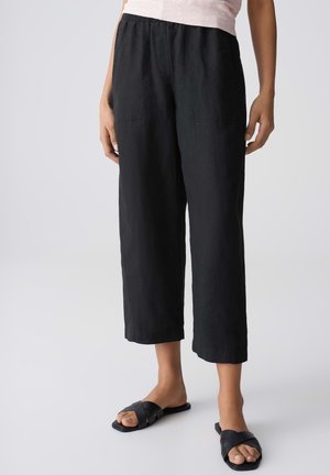 Personne portant un pantalon court noir ample et des sandales noires à enfiler, debout devant un fond clair uni.