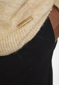 Pull en maille beige avec une texture côtelée, agrémenté d'une étiquette en métal doré, superposé à un tissu foncé.