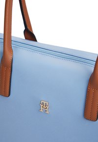 Φωτεινό γαλάζιο τσάντα χειρός με καφέ δερμάτινες λαβές και χρυσό λογότυπο "TH" με διαπλεκόμενα γράμματα κοντά στο πάνω φερμουάρ.