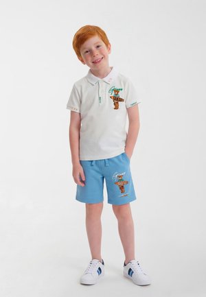 Garçon roux souriant portant un polo blanc et un short bleu avec un ours en peluche et le logo "Guess", debout avec une main dans la poche.