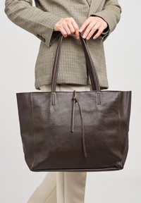 Borsa tote in pelle marrone scuro con finitura testurizzata, caratterizzata da due manici e un dettaglio centrale a cravatta, appoggiata su uno sfondo chiaro.