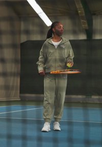Dvodijelni atletski komplet u zelenoj boji kadulje, koji se sastoji od dukserice s patentnim zatvaračem i odgovarajućih hlača. Drži tenis reketa i lopticu, stojeći na plavom terenu.