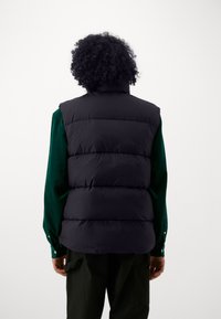 Gilet invernale nero con colletto alto e design trapuntato orizzontale, indossato sopra una camicia verde, abbinato a pantaloni cargo scuri. Hardware minimale.