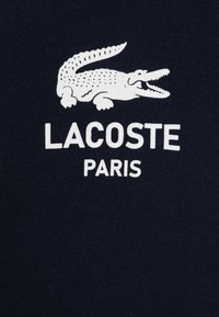 Camiseta de algodón azul marino con un logo de cocodrilo blanco y el texto "LACOSTE PARIS" debajo en letra negrita. Textura suave, diseño casual.
