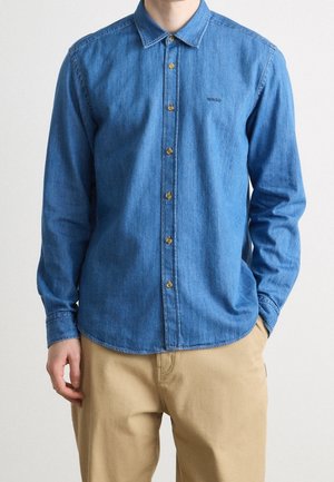Mand iført en langærmet blå denim skjorte med beige bukser, den ene hånd i lommerne og synligt "BOSS" logo på skjortens bryst.