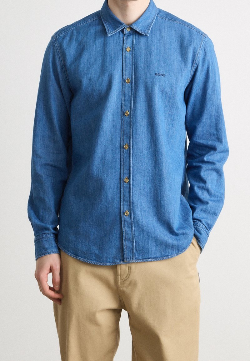 Homme portant une chemise en jean bleue à manches longues avec un pantalon beige, une main dans la poche, et le logo "BOSS" visible sur la poitrine de la chemise.