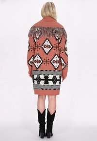 Gebreide jurk in terracotta met zwart-witte geometrische patronen, voorzien van een franjeshals en lange mouwen, afgewerkt boven de knie.