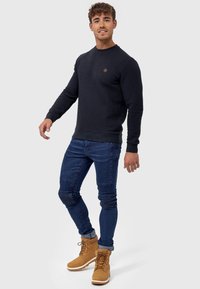 Navy textured sweatshirt with ribbed cuffs, worn with fitted blue jeans and tan lace-up boots. Features a small logo on the chest.