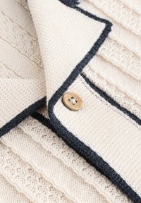 Strikket sweater i cremefarve med teksturerede striber, mørk navykant og en træknap som detalje.