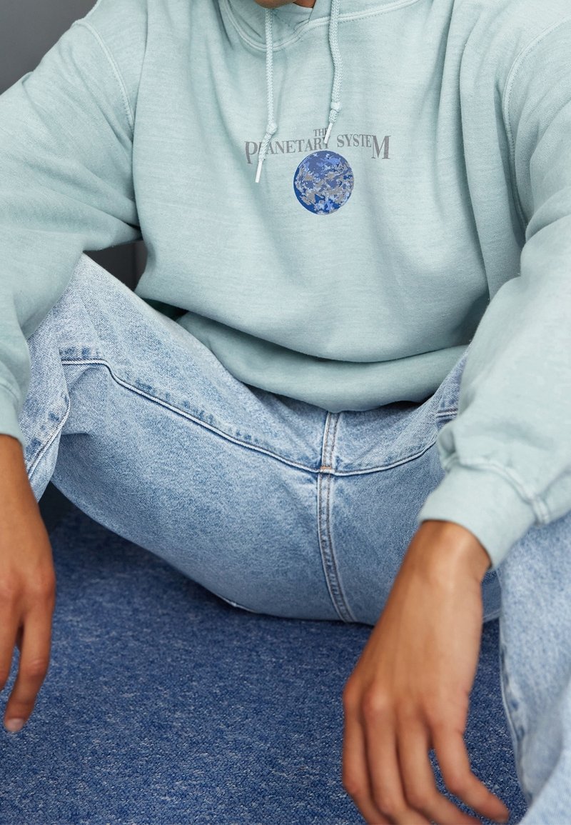 Ljusblå hoodie med en logotyp som föreställer jorden, kombinerad med slitna blå jeans. Tyget verkar mjukt med en avslappnad passform och snörningsdetalj.