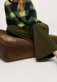 Sweter dziany w kratę w kolorach zielonym, niebieskim i żółtym, zestawiony z oliwkowozielonymi szerokimi spodniami, siedzący na brązowej poduszce w prążki.