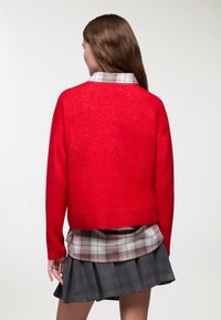 Czerwony sweter z dzianiny o skróconym fasonie, z teksturowanym wykończeniem. Połączony z koszulą w kratę i szaro plisowaną spódnicą.