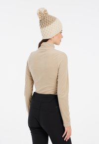 Maglione beige in pile a maniche lunghe con collo alto, abbinato a pantaloni neri a vita alta. Un berretto lavorato a maglia crema e marrone chiaro ha un pom-pom sopra.