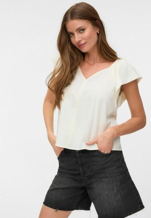 Jeune femme aux longs cheveux bruns portant un haut blanc à manches courtes et col en V ainsi qu'un short en jean noir taille haute, les mains dans les poches.