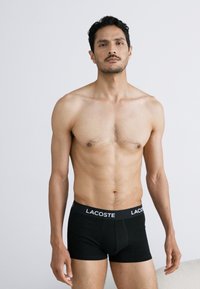Schwarze Baumwoll-Boxershorts mit einem weißen Lacoste-Logo auf dem Bund. Das Modell hat eine schlanke Figur und steht vor einem neutralen Hintergrund.